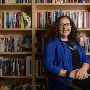 Woman with curly brown hair and blue jacket smiling at the camera and sitting in front of a book shelf