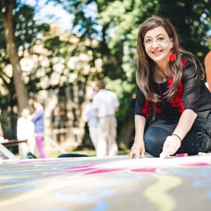 A woman with long brown hair draws in colored chalk outdoors on a sunny day