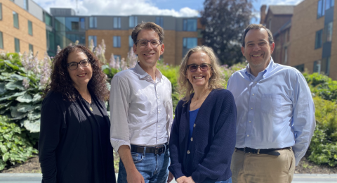 Four people stand smiling in front of the camera with greenery in the background