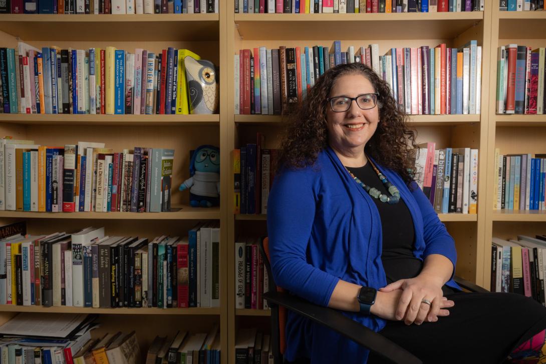Woman with curly brown hair and blue jacket smiling at the camera and sitting in front of a book shelf