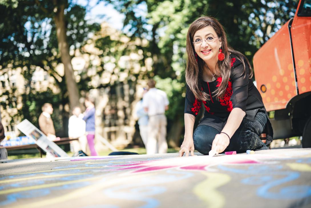 A woman with long brown hair draws in colored chalk outdoors on a sunny day